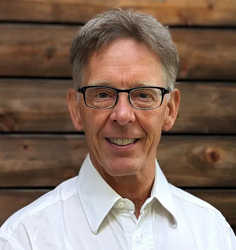 Mann mit grauen Haaren und Brille trägt ein weißes Hemd vor einer Holzwand.